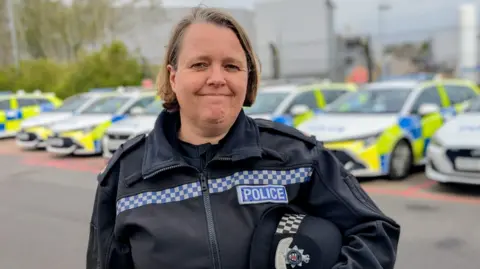 Supt Karen Corrigan wearing a black police uniform and holding a black hat. She is standing in a car park, and in the out of focus background, you can see a row of around seven parked police cars with blue and yellow hi-vis decals.