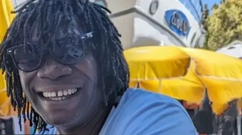 Image of Derek Thomas, a man with dark dreadlocks wearing a blue t-shirt and sunglasses
