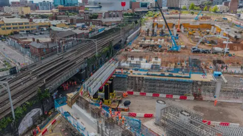HS2 Limited An aerial image including rail lines going from the bottom left to the top of the photo in the middle. The two silver beams are to the right of the tracks. Cranes are in the top right of the image and buildings are in the distance.