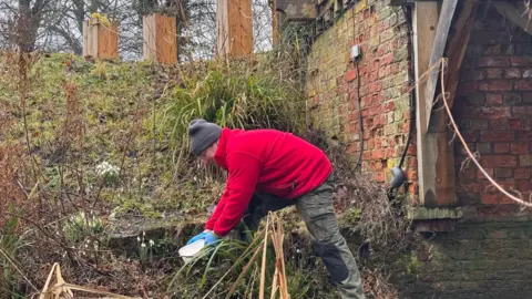 National Trust A man in an anorak and woolly hat, is bending over holding a box outdoors, on the banks of a moat 