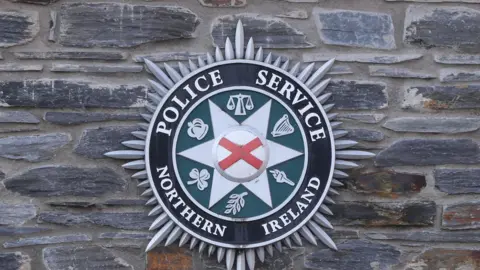The logo of the Police Service of Northern Ireland is mounted on a stone wall at a police station. The name of the force surround the crescent. 