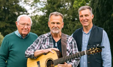 Three men with short grey hair stand in a garden in front of trees. The man in the middle has a guitar strapped over his shoulder.