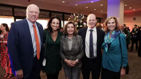 Department for Communities Five well-dressed adults, three women and two men, stand close together, smiling at the camera at an indoor evening event. They are in a warmly lit reception space with other guests mingling in the background.