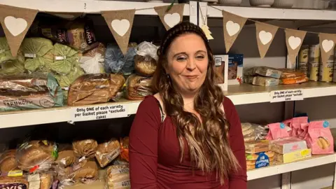 Lianne Simpson smiles and stands in front of shelves of bread and cakes. Hessian bunting with white love hearts are above her head and hang from the shelving. 
