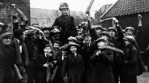 A black and white photo of several young boys in caps and suits waving rolls of leather about three foot long above them. In the middle you can see an old man wearing a garlanded top hat being held up by three other men who are shouting with their mouths wide open, also wearing garlanded top hats. Brick buildings can be seen behind them.