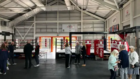 A boxing gym with two boxing rings in the background next to the entrance, and punching bags lined up on the right. People are milling around the floor.