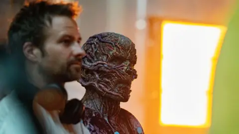 Netflix Vecna, played by Jamie Campbell Bower is in character as Vecna. He is wearing a prosthetic face mask which has vine-like threads. He is standing beside co-creator Ross Duffer.