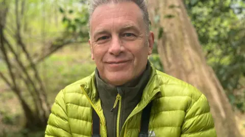 Fiona Irving/BBC Chris Packham, chief ecologist for Ecotalk, pictured at Bowyers Wood, East Sussex. Chris is wearing a lime green jacket and dark green fleece. He has a pair of binoculars around his neck. Chris is smiling to camera and has grey hair.
