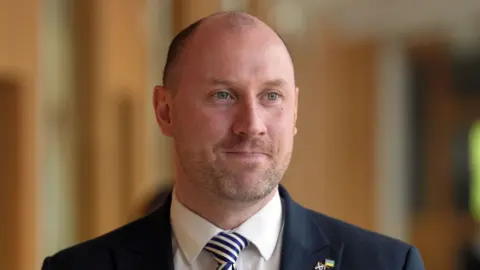 PA Media Neil Gray standing in a corridor at the Scottish Parliament building with a smile on his face. He is wearing a dark jacket, a blue and white tie and white shirt.