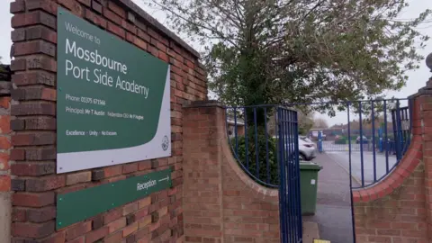 The entrance to Mossbourne Port Side Academy, showing a green sign on a brick wall and blue metal gates leading into the school grounds.