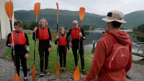 A dad and his three children - a girl and two older boys, stand in a line in water safety gear as an instructor gives a safety talk at the side of the loch. They are all holding bright orange paddles 