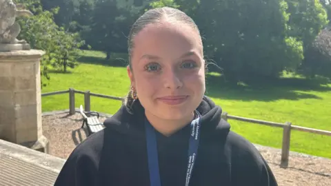 A young girl who is wearing a black hoodie and has her hair tied back looks at the camera and smiles. In the background, you can see a bench and some greenery which is part of the Singleton campus at Swansea University.