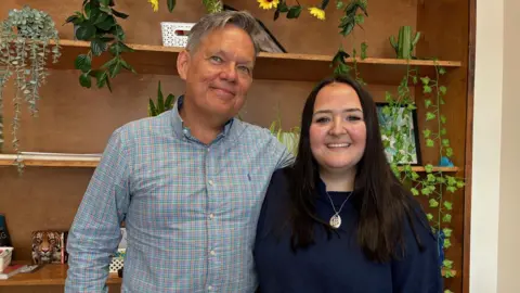 The image shows James standing with his arm around his daughter Imogen. They are both smiling.