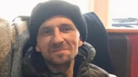 A slightly grainy photo of a man wearing a beanie hat and coat sitting in a chair. He appears to have some beard stubble and is smiling at the camera