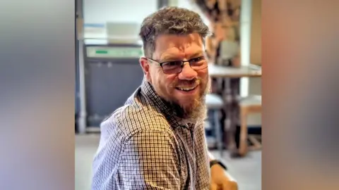 Jason Tholen is sitting and is in the middle of the image. He has short brown hair and a medium length brown beard. He is wearing a blue and red tartan shirt.