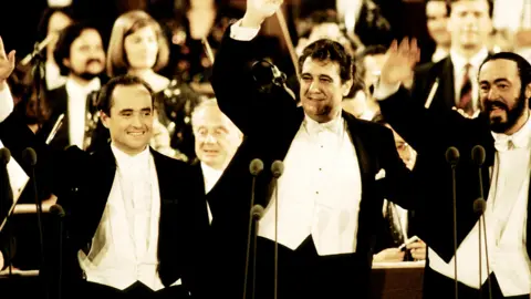 Getty Images Jose Carreras is on the left of the photo wearing white tie and tails. He has slightly receding dark hair and eyes and has his right arm raised in a wave. He is smiling and looking to the right of the camera. Placido Domingo is the tallest of the three in the middle. He has thick brown hair and is smiling. He is dressed the same and has the same pose but looks to the left. Luciano Pavarotti on the right has dark hair and a beard and is smiling to the left of the photo. He also wears tails. There are three microphones in front of each man. Behind them members of an orchestra are also standing but are out of focus.