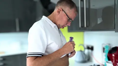 Marc Powell is wearing a white zip-up polo shirt and is in the kitchen looking at a feeding tube. 