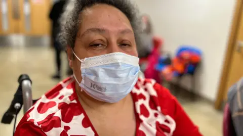 A woman in a wheelchair at an urgent treatment centre. She has greying dark hair and is wearing a surgical mask, and a red and white top.