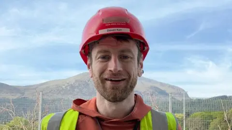 Mr Kerr has dark brown hair and facial hair he is smiling at the camera as he wears a red hard hat, a red hoodie and a yellow hi-vis. Behind him is a mountain range and a metal fence.