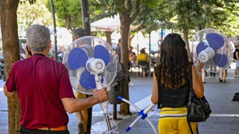 Two people seen from behind carrying domestic electric fans down a street