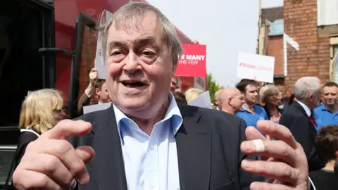 John Prescott out campaigning in Hull in 2017 with Labour Party supporters in the background. He is wearing a suit with a pale blue shirt and gesturing at the camera, he has a plaster on one finger. 