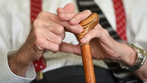 An elderly man holding a walking stick. The image is a close up of his hands. 
He is wearing a watch on this left wrist and a white shirt with red braces and a tie with black trousers.