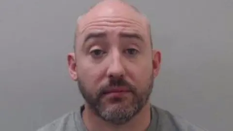 A man with a dark and grey beard looks into the camera. He is bald and is wearing a grey sweatshirt.