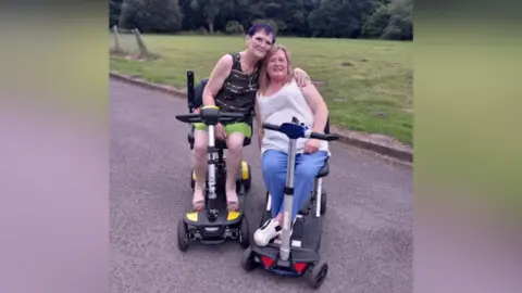 Christine, who wears green shorts and a black vest top, and Heather, who wears blue trousers and a white west top, both sit on mobility scooters in a park.