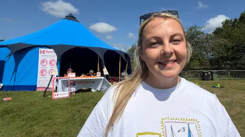 BBC/Jim Scott Charlotte Graham, wearing a white shirt with a picture frame design, stands in front of the blue sensory tent which has been installed at the rear of the festival and away from the noise.