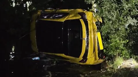 A yellow car can be seen on its side in the river, while an emergency worker in yellow high-viz is too the right with a green rucksack.