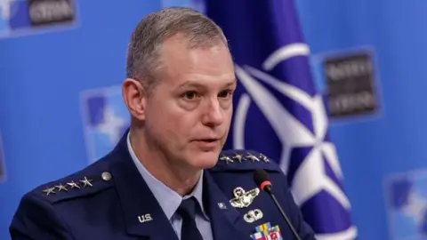 A clean-shaven man in a blue military uniform sits at a table in front of a Nato flag for a press briefing.