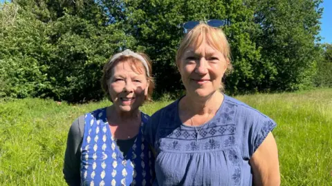 Ellen Knight/BBC Lynne and Nikki stood side by side in the wild grass meadow. They are both wearing blue blouses, and Nikki has a pair of sunglasses on her head. The grass around them is long and green, and there is a row of trees behind them. The sky is blue and there is strong sunshine. 