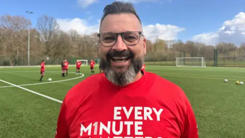 A man with short grey/brown hair and a beard and wearing glasses is smiling as he stands beside a football pitch. He is wearing a red Every Minute Matters top.
