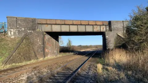 A bridge that is over a railway line between Bristol and Taunton. There are two tracks underneath the bridge, with hedging to the right. The bridge has a stone structure with a metal footway.