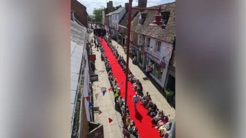 Shot from a second-floor window showing pedestrianised shopping street with red carpet running down the middle. The carpet has chairs along both sides in which spectators are sitting. There is a black stage at the top end. A man is walking down the carpet showing off an outfit. There is red, white and blue bunting stretched out above the street.