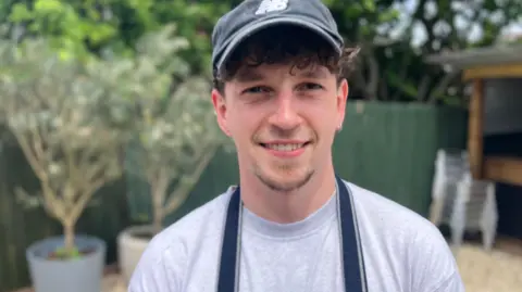 A close-up photo of Zack shows wearing a dark baseball cap and a striped apron over a light-coloured T-shirt, standing outdoors in a cafe. 