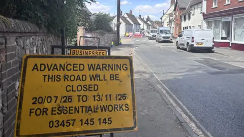 Matt Marvel/BBC A sign beside a road that reads: "Advanced warning. This road will be closed 20/07/26 to 13/11/26 for essential works." Cars are parked along the opposite side of the road. The road is lined by buildings on the right and a brick wall on the left. 
