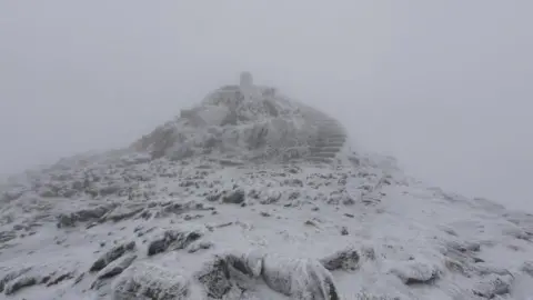The summit of Yr Wyddfa covered in snow and ice. There is poor visibility all around.