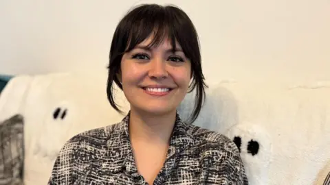 Natasha Molyneux-Smith is smiling, and has dark hair loosely tied up and a fringe. She is wearing a black and white patterned shirt and sitting on a sofa with a cream throw over it.