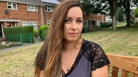 A woman with long brown hair, swept to one side, sat on a bench. She is wearing a short sleeved top with black lace shoulders and purple front