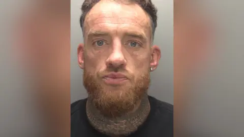 Close up photo of a man with short brown hair and a beard. He has an earring, a neck tattoo and is wearing a black top.