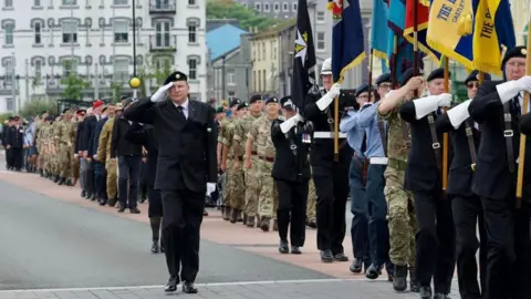PhotoMann Armed forces personnel in uniform lined up standing to attention. A senior official salutes while standard bearers hold the standards upright.