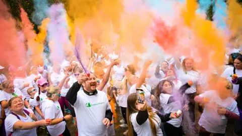Gateshead Council Runners taking part in a Pride 5k run. They are wearing white T-shirts and are throwing colourful powdered paint into the air.