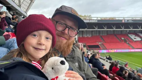 Lucy Roberts Willow and her dad Matt hug and smile into the camera in the stands at a Wales game. Willow is wearing a red woolly hat and cuddling a white teddy bear. Her dad Matt has glasses and a beard, and is wearing a grey raincoat and cap.