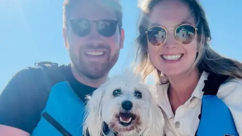 Joe McKenna Joe (left) and Charlotte (right) smile at the camera with Max in the centre. They are all smiling at the camera. Joe and Charlotte are both wearing sunglasses and blue lifejacket vests. 