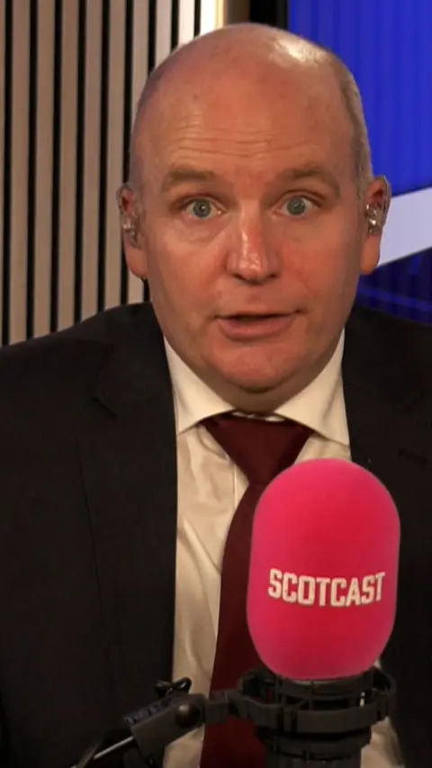 Glenn Campbell talks into a studio radio mic. he is wearing a black suit jacket, white shirt and a tie.