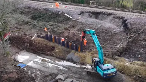 A work site with a blue digger and people in orange hi vis suits. A railway line sits on an embankment where a large part of the land has given way in a landslip.