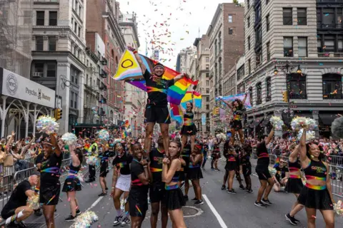 Alex Kent/Getty Images New Yorkers celebrate during the annual Pride March into the West Village on June 30, 2024 in New York City.  The images shows cheer leaders performing with sparkling pom poms while walking down a street with New York city skyline in the background