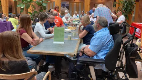 A table with around 20 people sat around it, some of whom are wearing orange t-shirts which say 'MND Association' on it.