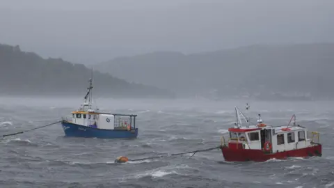 Two small creel boats, one white and blue and the other white and red, strain at their moorings at Fort William during August's Storm Floris.